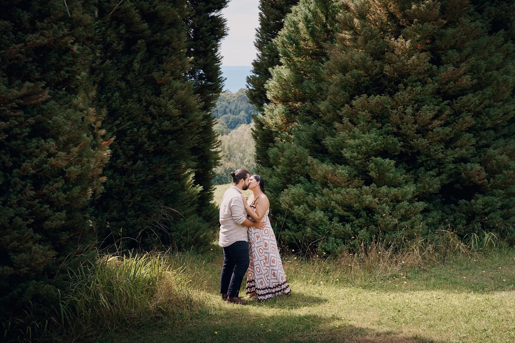 A couple sharing a kiss in a lush green outdoor setting with tall trees on either side.