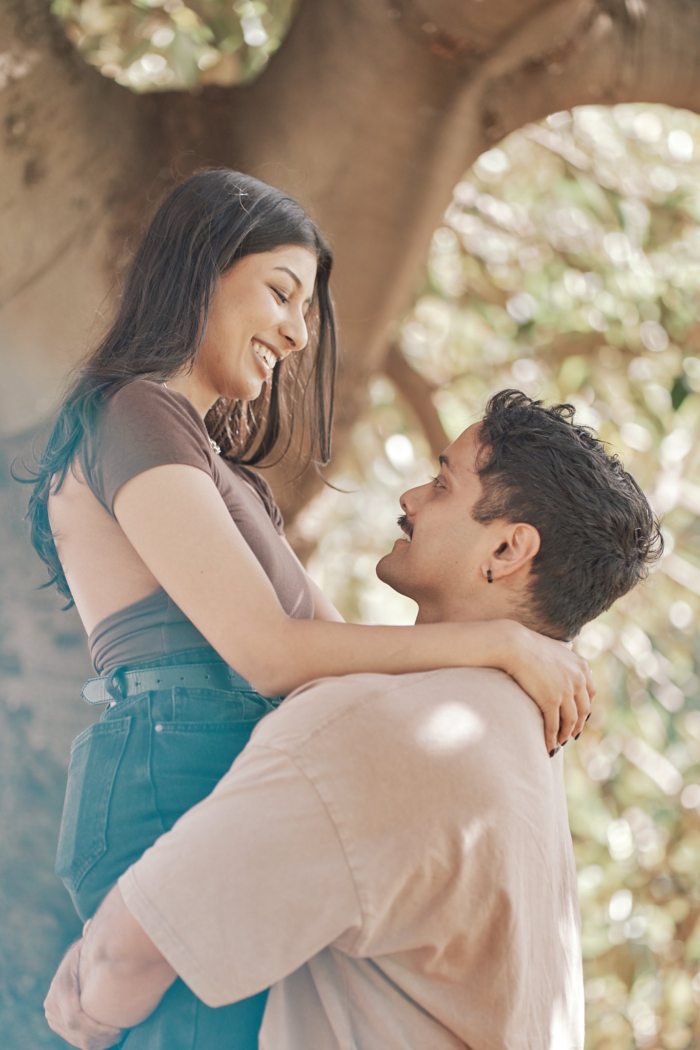 A man holding a woman while she smiles and looks at him outdoors near a large tree with sunlight filtering through the leaves.