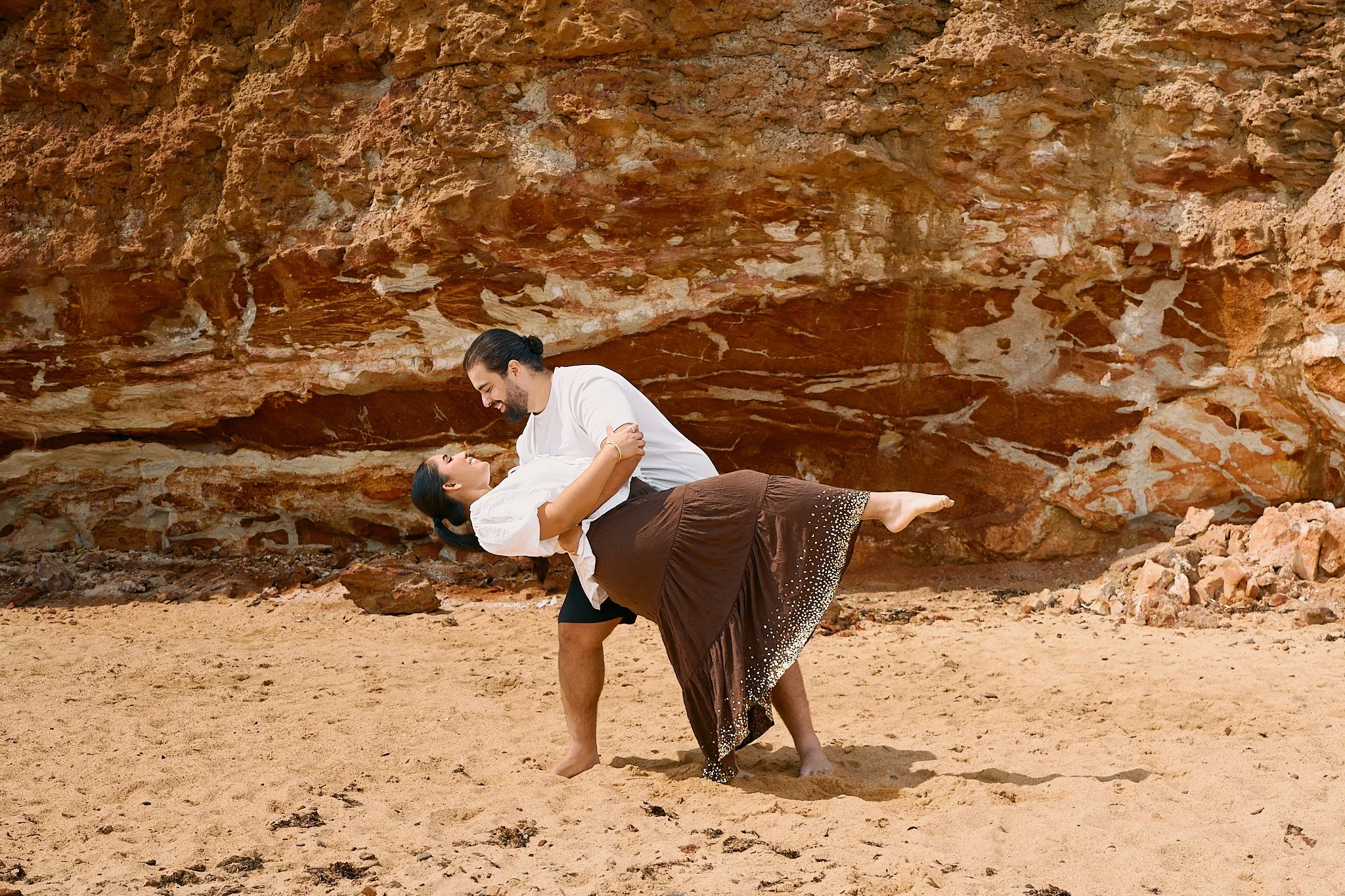 A man and woman are dancing on a sandy beach with large rocks and reddish cliffs in the background. The man is holding the woman in a dip pose, smiling at each other.