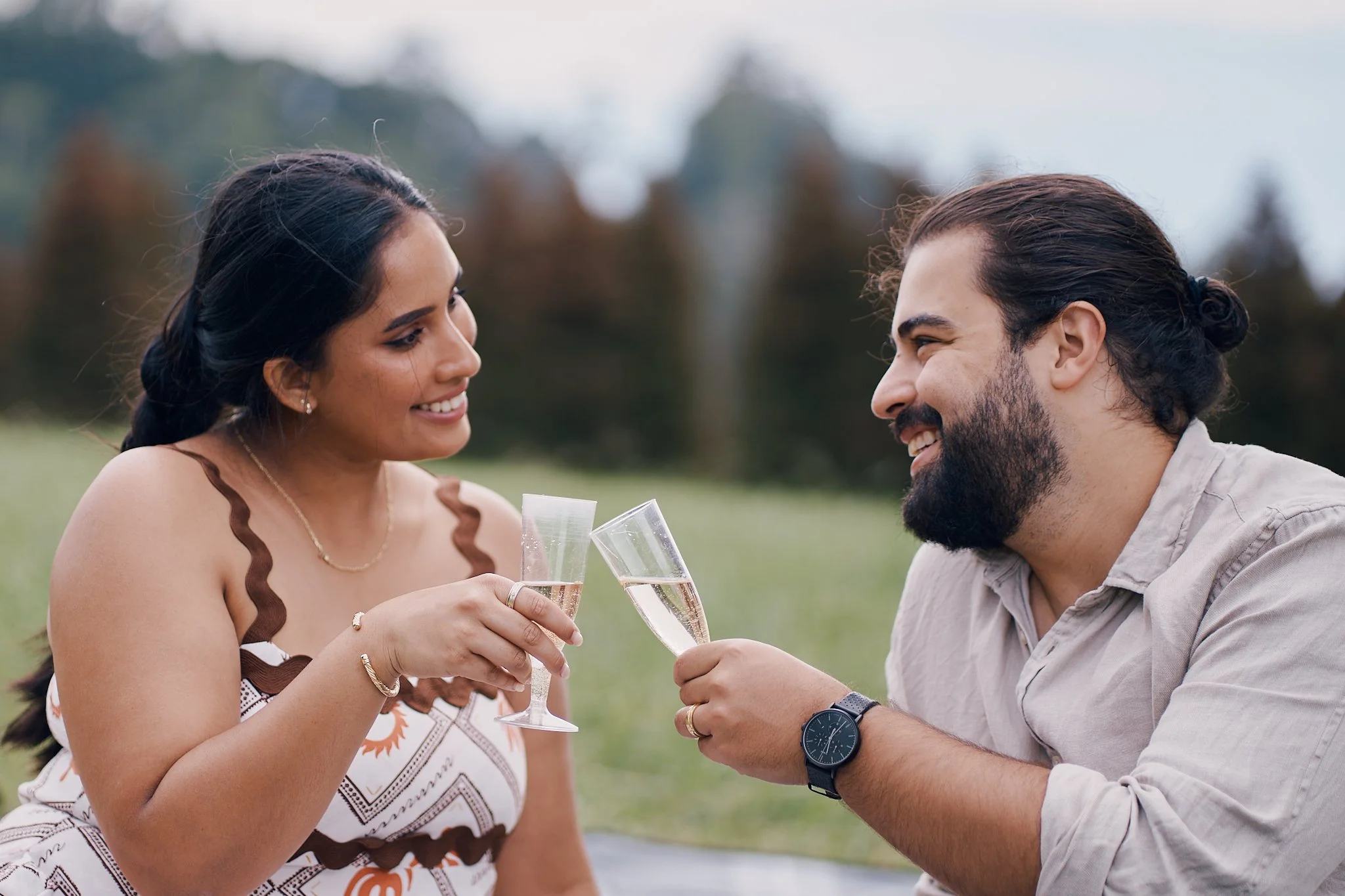 A man and woman clinking glasses of champagne outdoors during daytime, smiling at each other with a scenic background of trees and a cloudy sky.