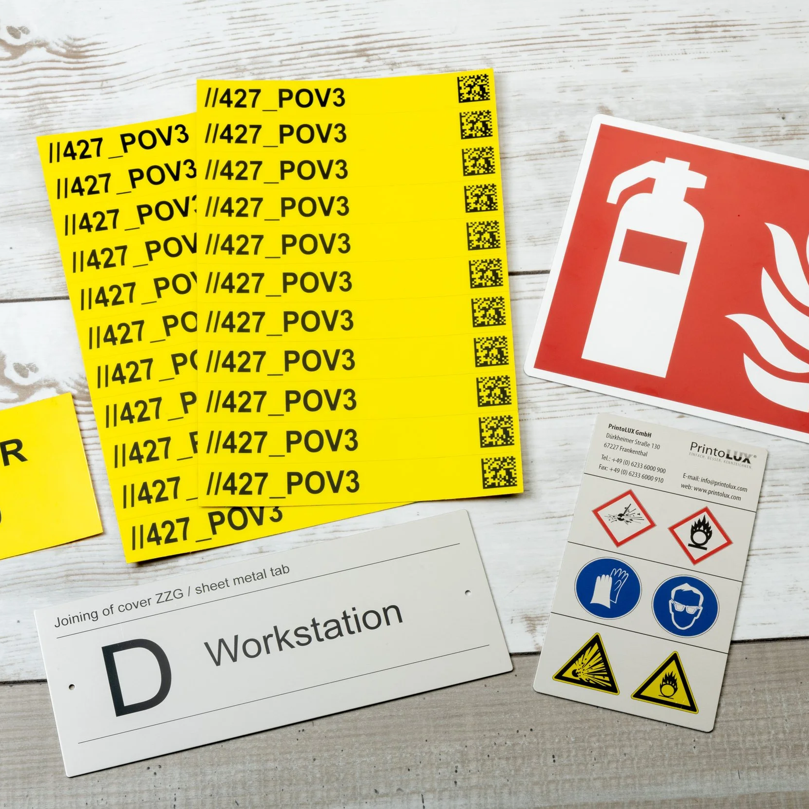 Multiple yellow tickets with black text and QR codes, a red fire safety sign, a business card, and a sheet metal work station label on a white wooden surface.