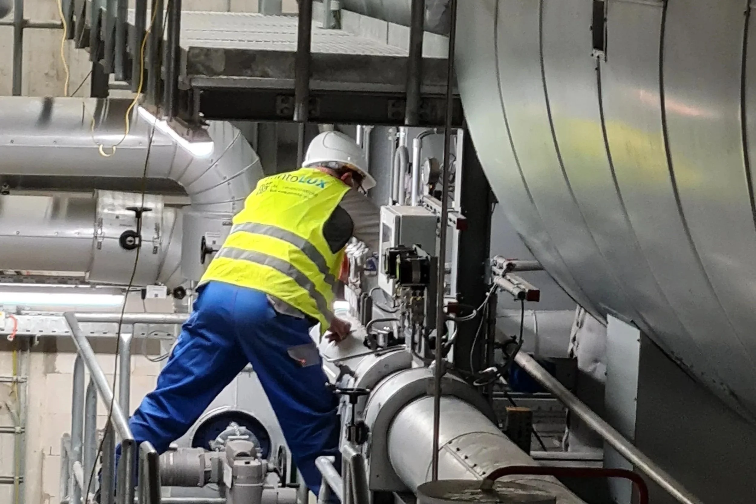 A worker in a yellow safety vest, blue pants, and a white hard hat working on machinery in an industrial setting, surrounded by large metallic pipes.
