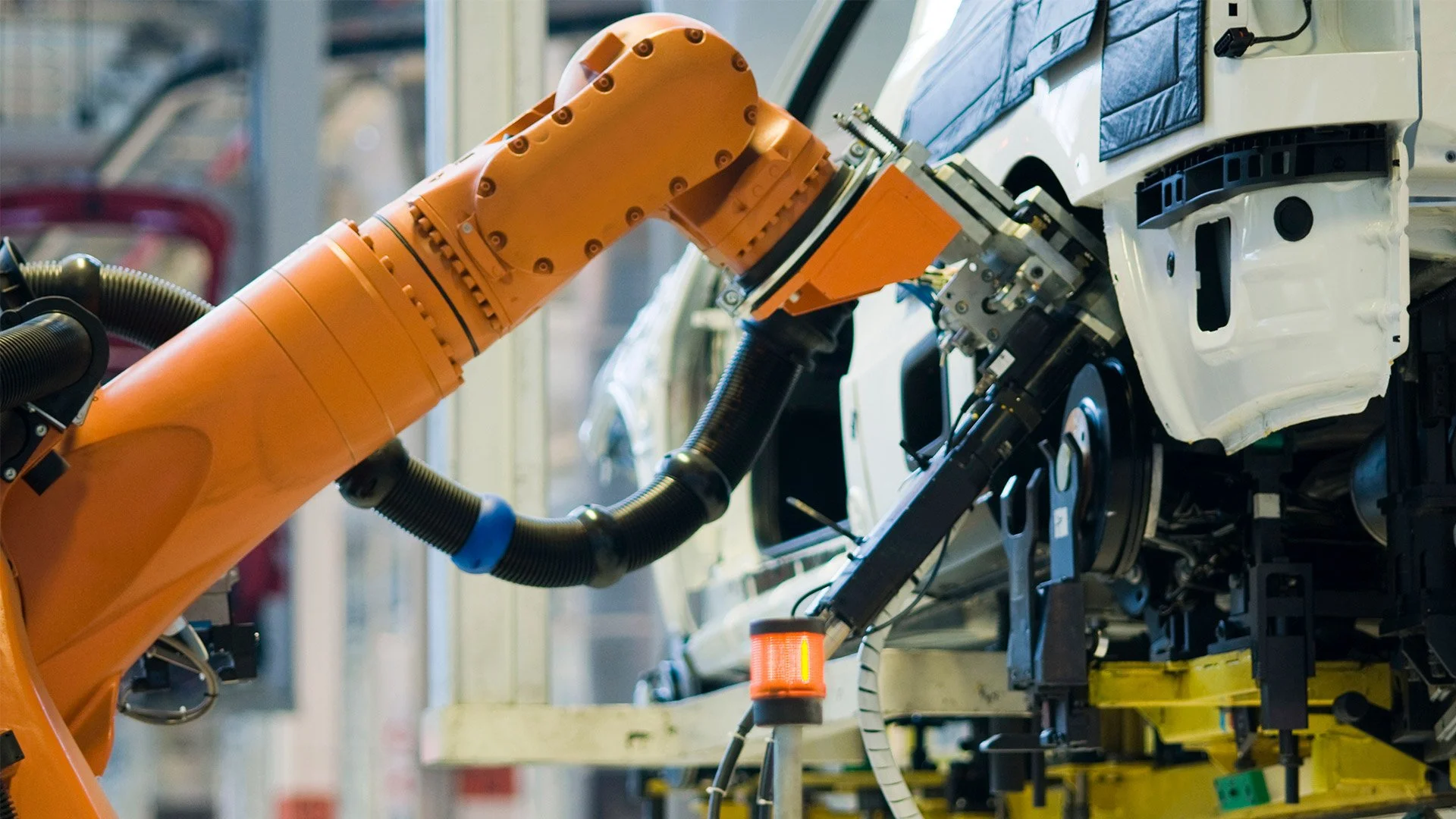 An orange robotic arm working on assembling or repairing a white vehicle in an automotive manufacturing plant.