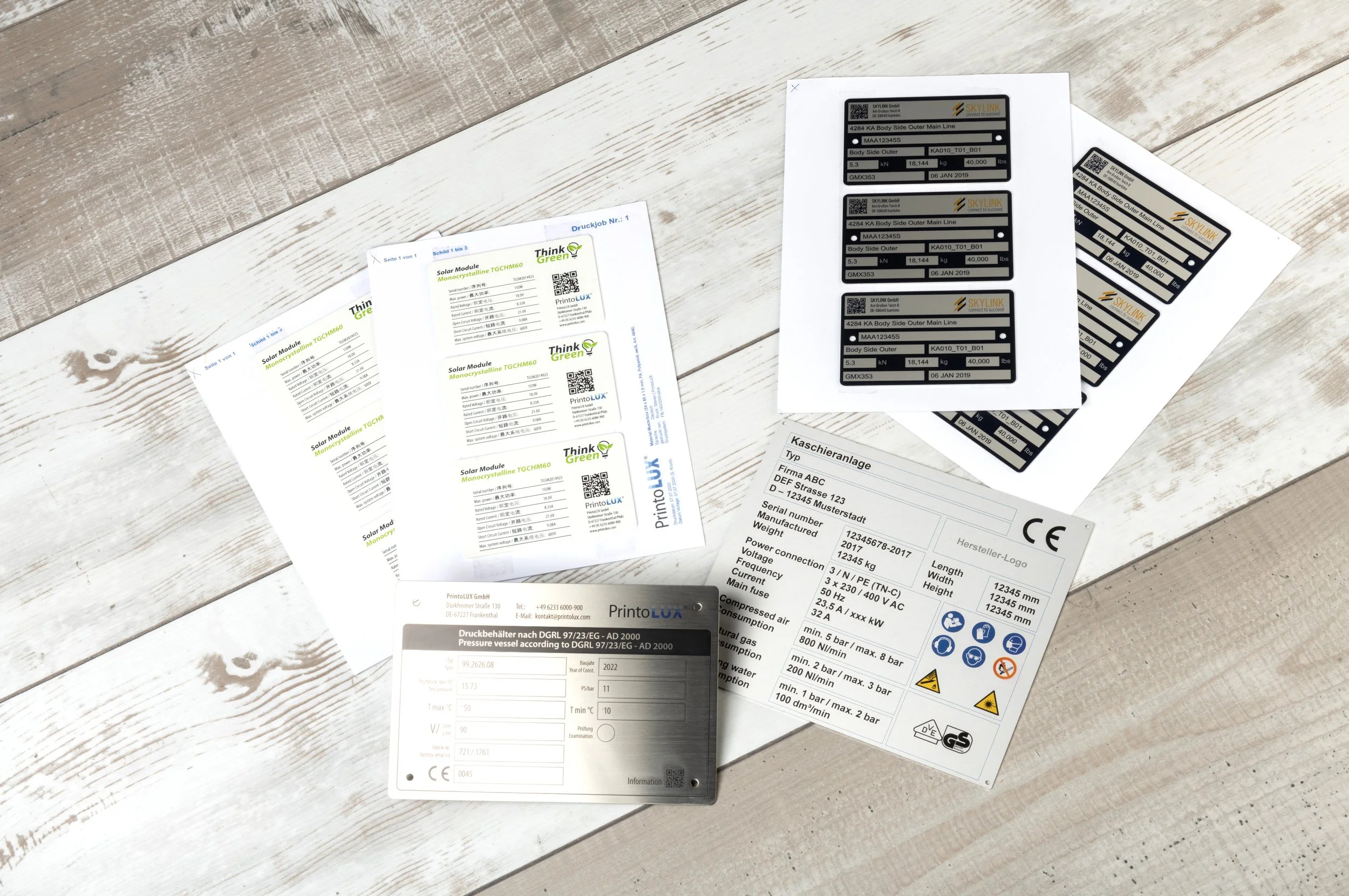 Several sheets of printed printolux labels and documents with technical information and barcodes on a white wooden surface.