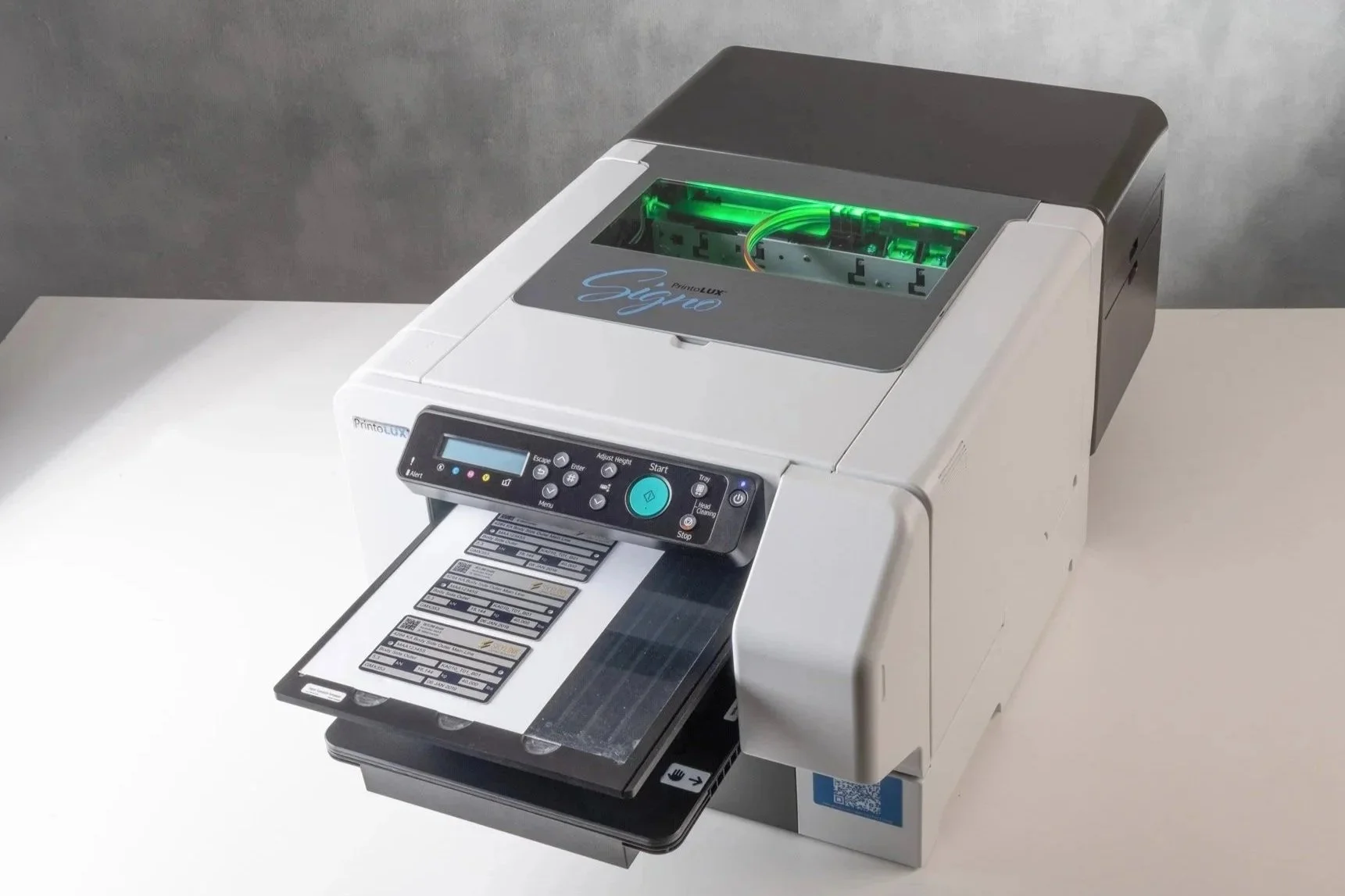 The Printolux Signo positioned on a white surface against a gray background. It is primarily white with a black and gray top section, and features a digital display panel and a tray with label plates.