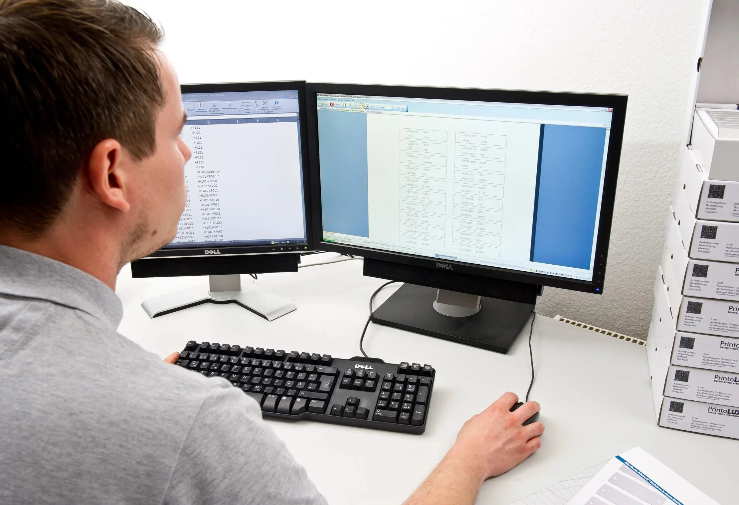A man working at a desk with dual computer monitors displaying spreadsheets, with a stack of Printolux materials on the side.