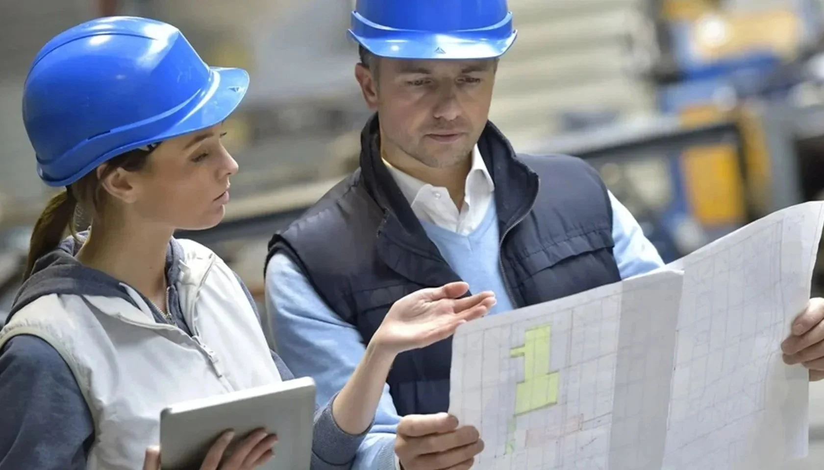 Two workers in a factory, both wearing blue safety helmets, are looking at a large blueprint or plan. The woman is holding a tablet, and the man is pointing at the blueprint.
