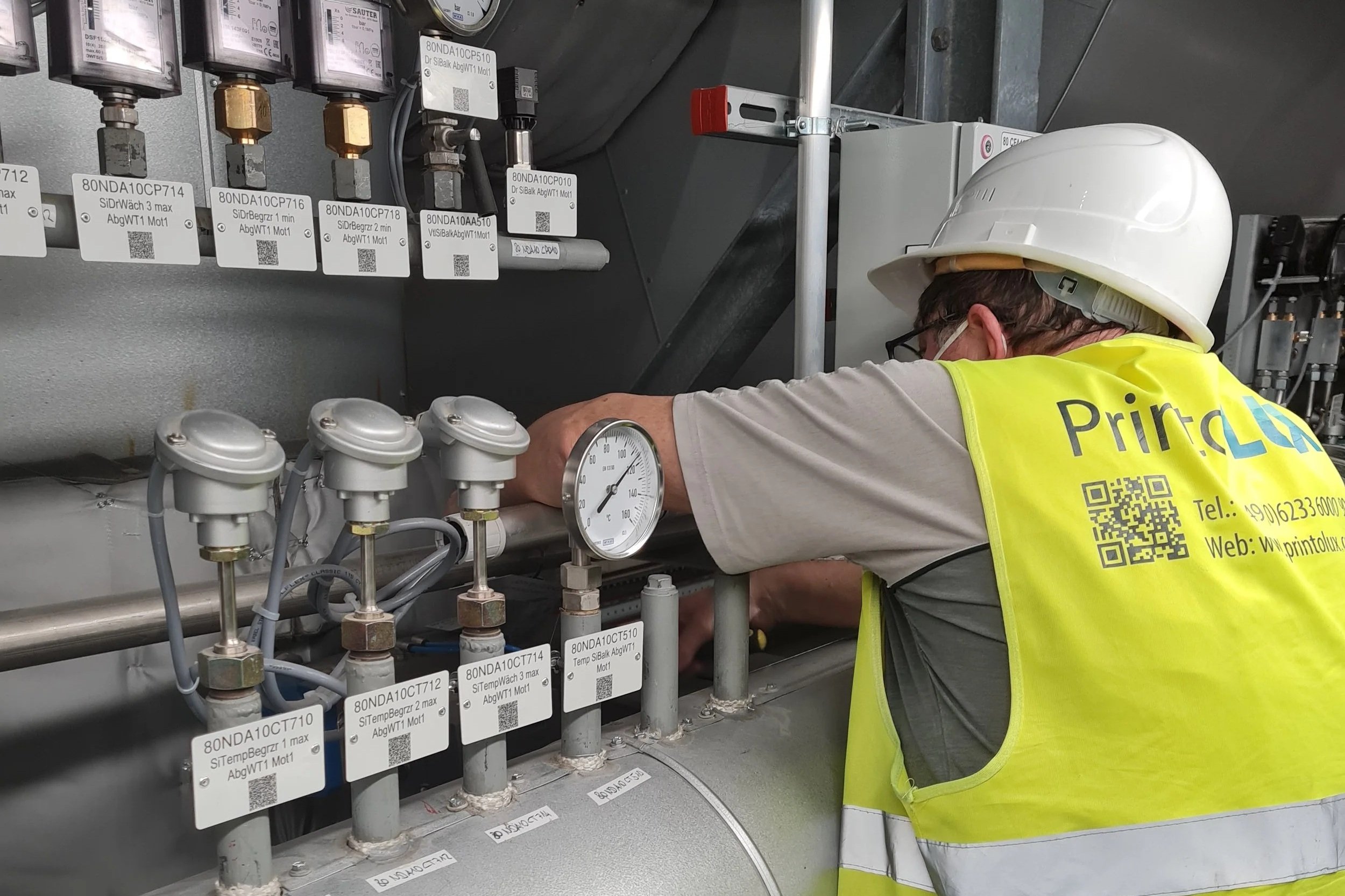 A worker wearing a yellow safety vest with a QR code and website, a white hard hat, and glasses is fixing or inspecting industrial equipment with gauges, pipes, and labels in a mechanical room.