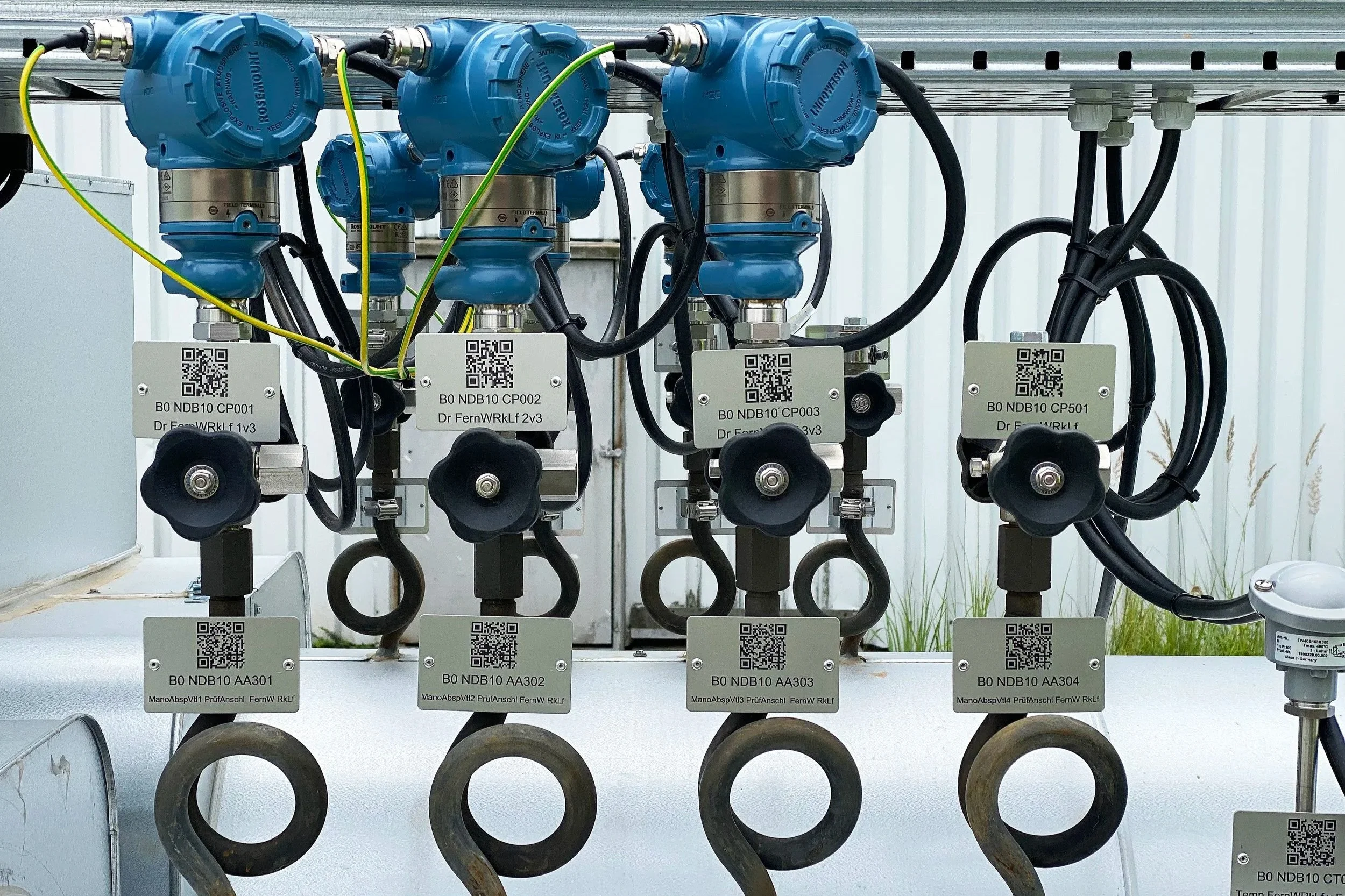 Industrial equipment with blue pneumatic actuators and black control valves, labeled with QR codes and part numbers, mounted on a metal surface with electrical wiring and pipes.