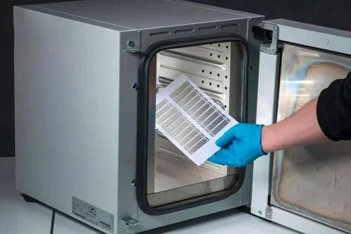 A person wearing a blue glove placing the small printed metal cable tags into a desktop size industrial oven for curing.