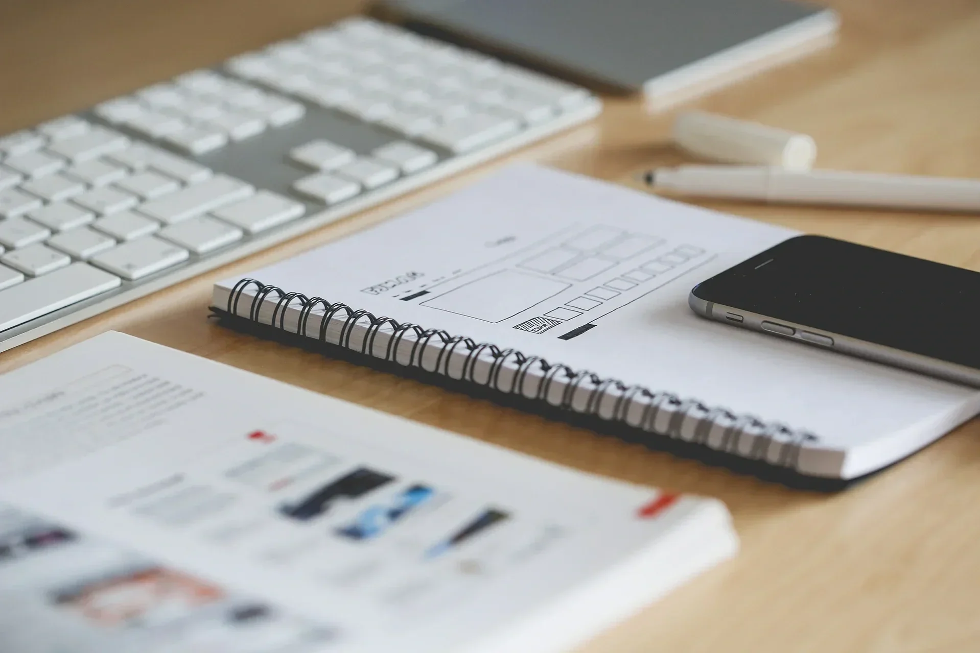 A desk with a computer keyboard, a white pen, a smartphone, and a notebook with sketches.