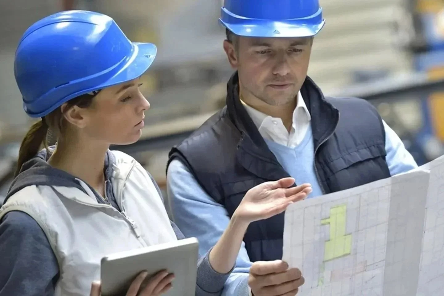 Two workers in a warehouse wearing blue safety helmets and discussing a large blueprint.