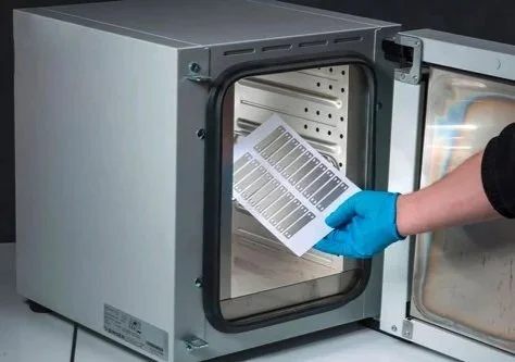 A person wearing a blue glove placing the small printed metal cable tags into a desktop size industrial oven for curing.
