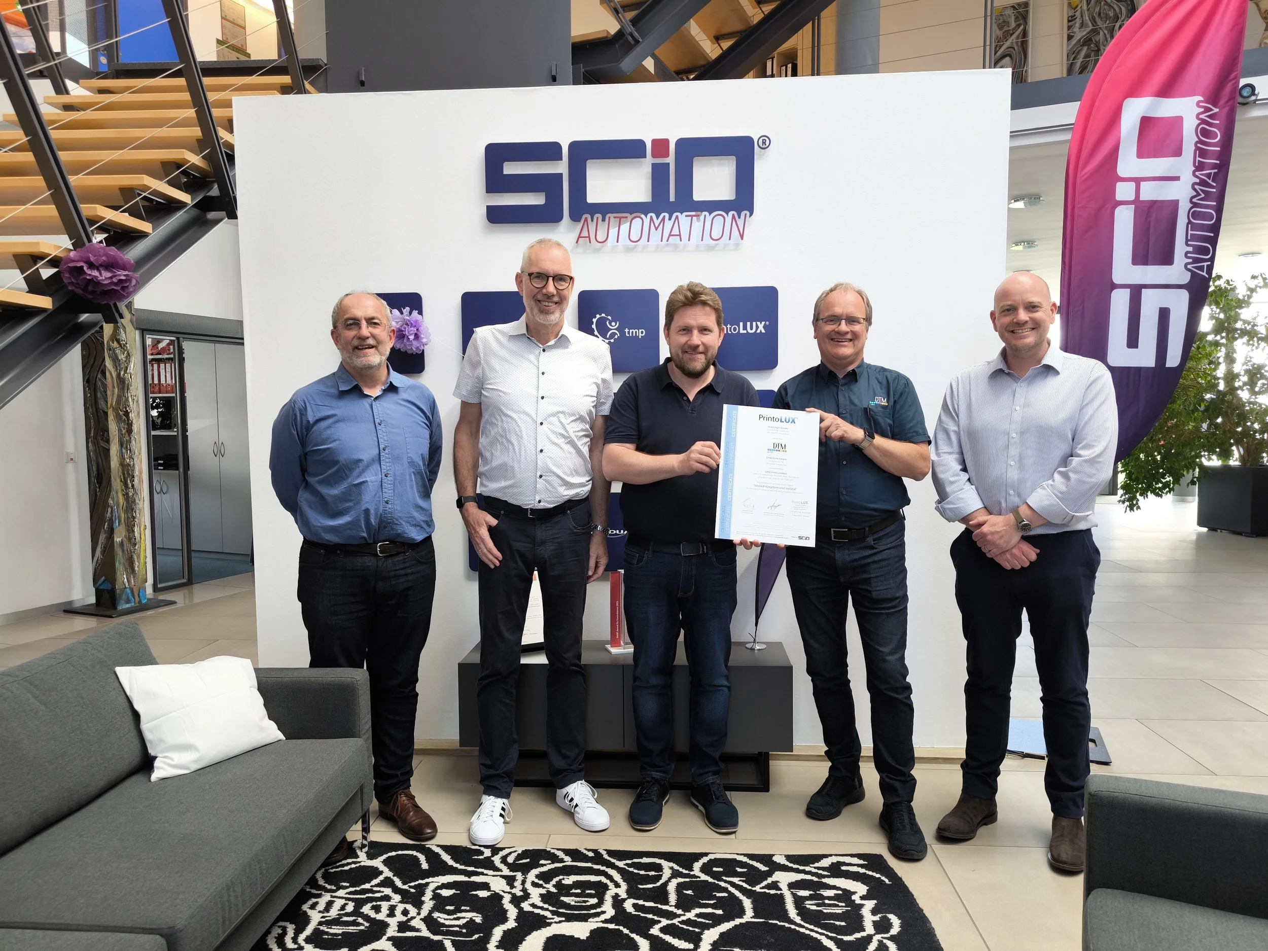 Five men standing in a row indoors, with a large sign behind them reading 'SCIO Automation' and a purple banner on the right. One man is holding a document or certificate, and they are smiling at the camera.