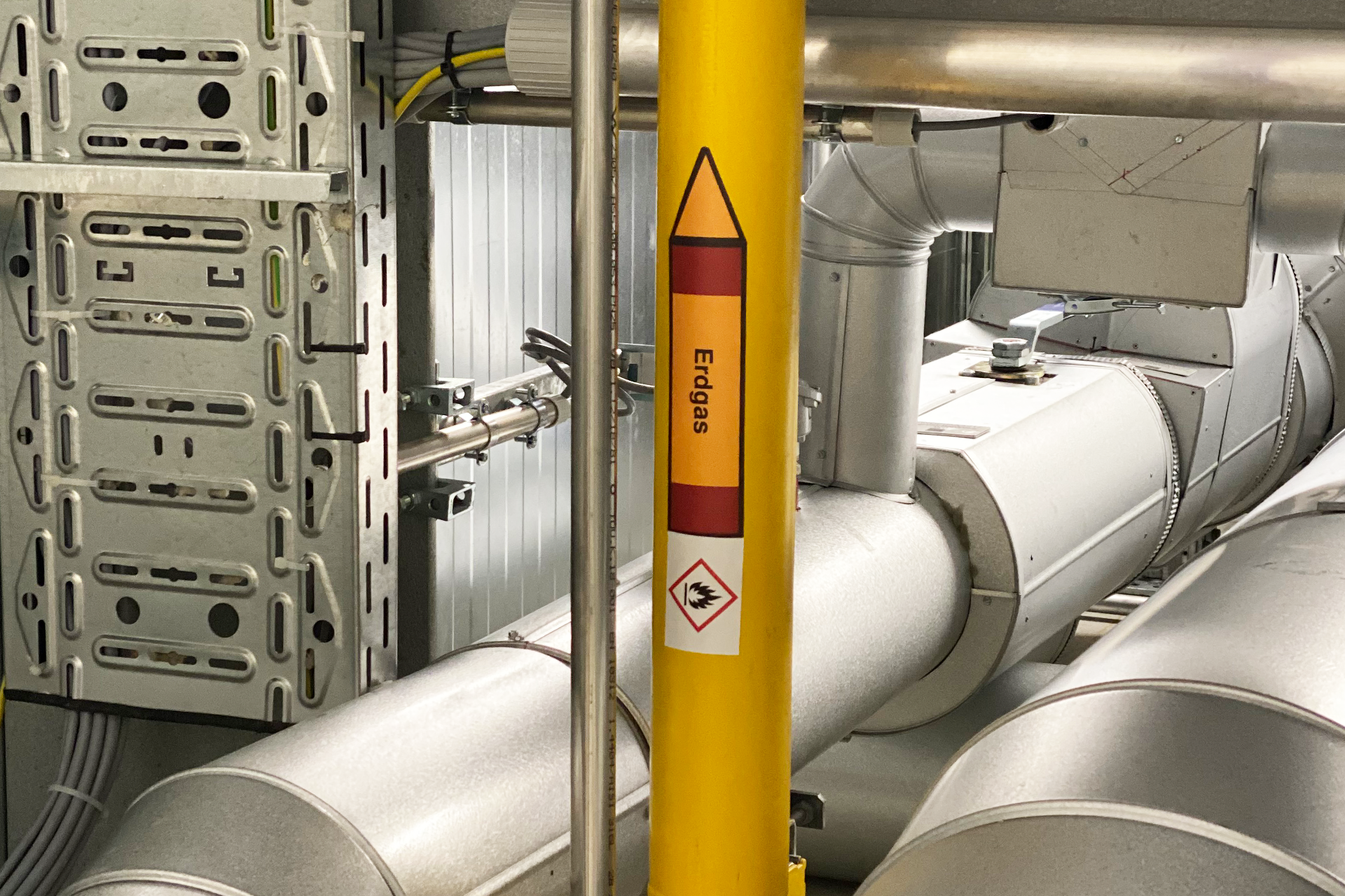 An industrial pipeline system with a PrintoLUX yellow hazard adhesive label indicating flammability, pointing upwards with the word "Erdgas" (natural gas), and a small warning symbol with a flame icon.