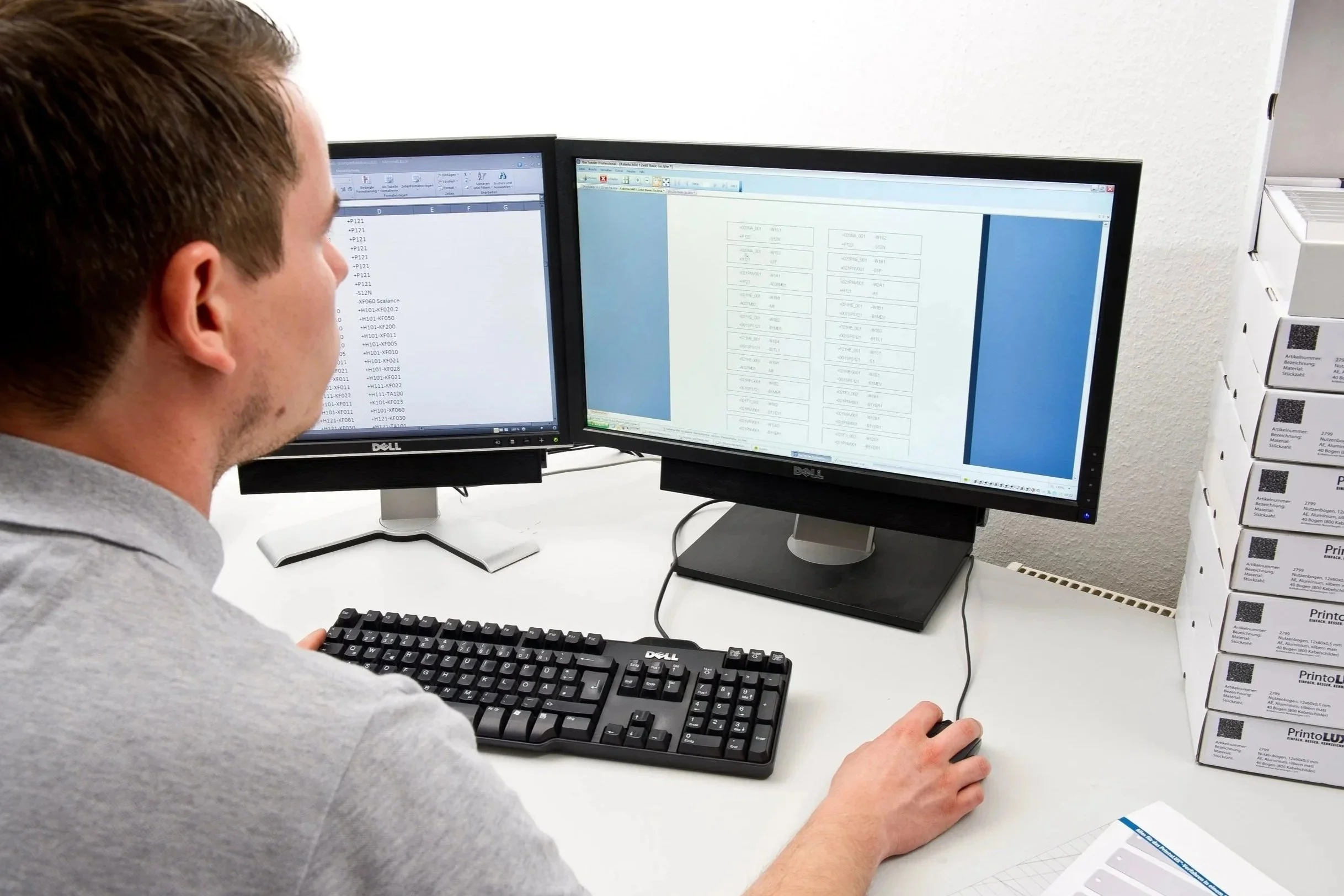 A man working at a desk with two monitors, typing on a keyboard and using a mouse, with stacked PrintoLUX material boxes on the right side of the desk.