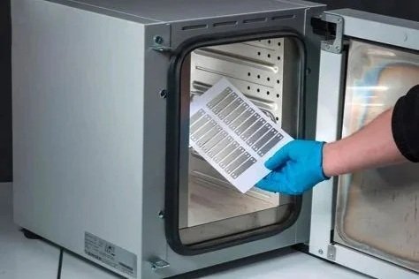 A person wearing a blue glove placing the printed sheet of metals tags into a small industrial oven for curing.