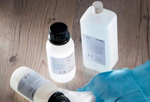 Cleaning supplies including a large white bottle, a smaller white bottle with a black lid, a large white bottle with a screw cap, a blue cloth, and a pair of white gloves on a wooden surface.