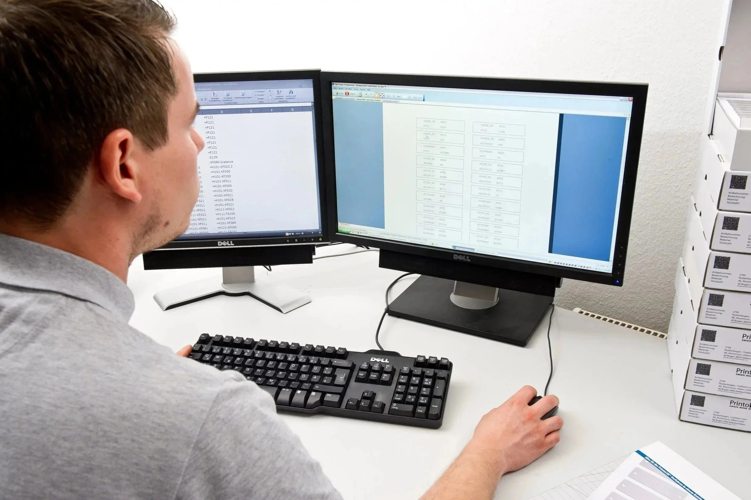 A man working at a desk with dual computer monitors displaying spreadsheets, with a stack of Printolux materials on the side.