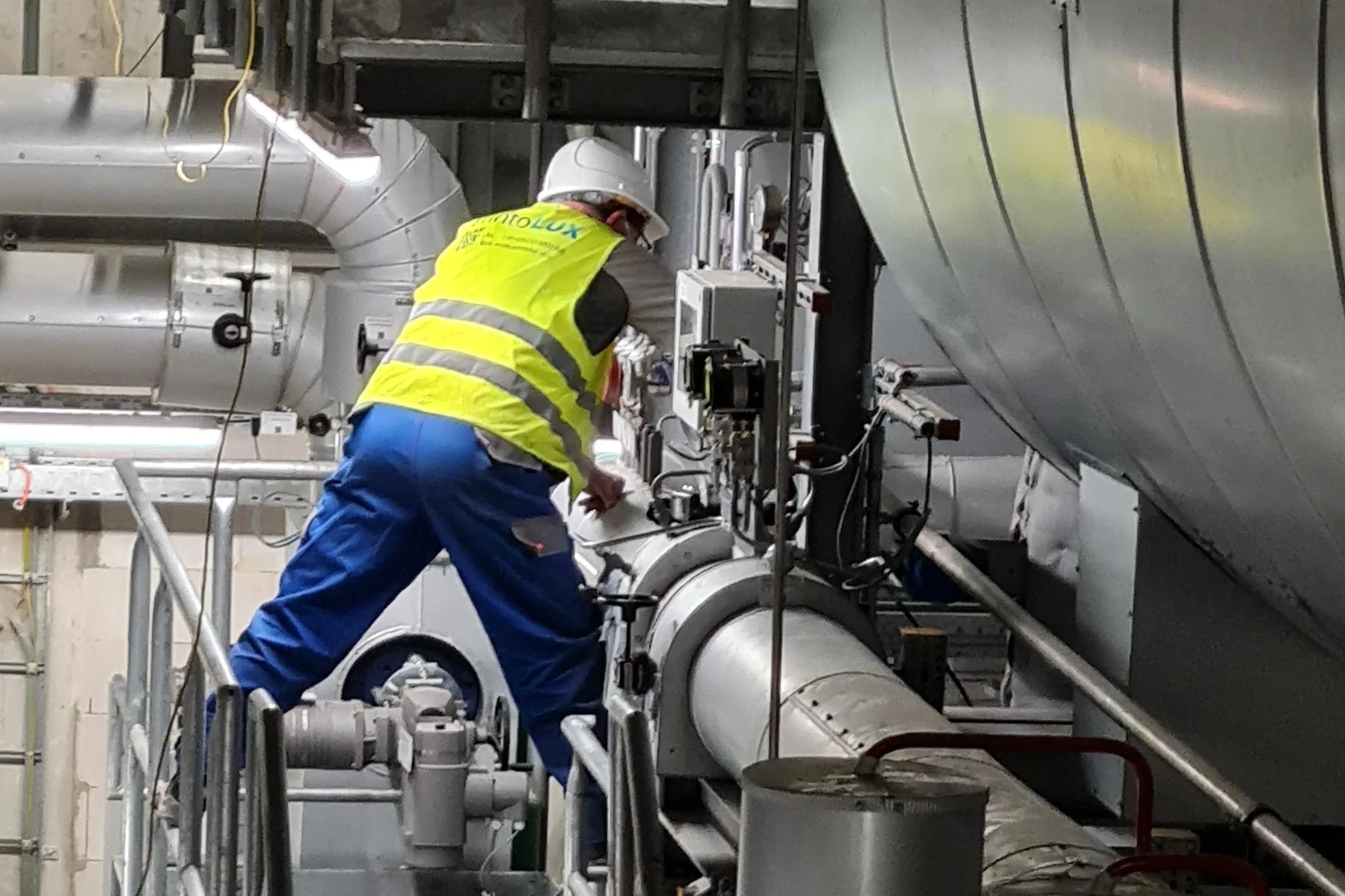 A worker wearing a white safety helmet, yellow safety vest, and blue work pants is working on industrial equipment in a factory or plant with large metallic pipes and machinery.