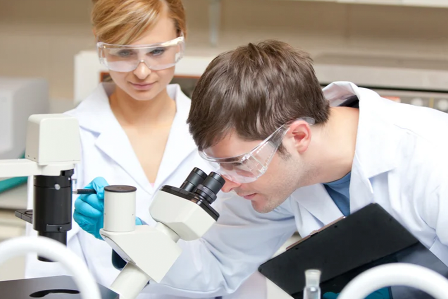 Two scientists in lab coats and safety goggles examining a sample through a microscope in a laboratory.