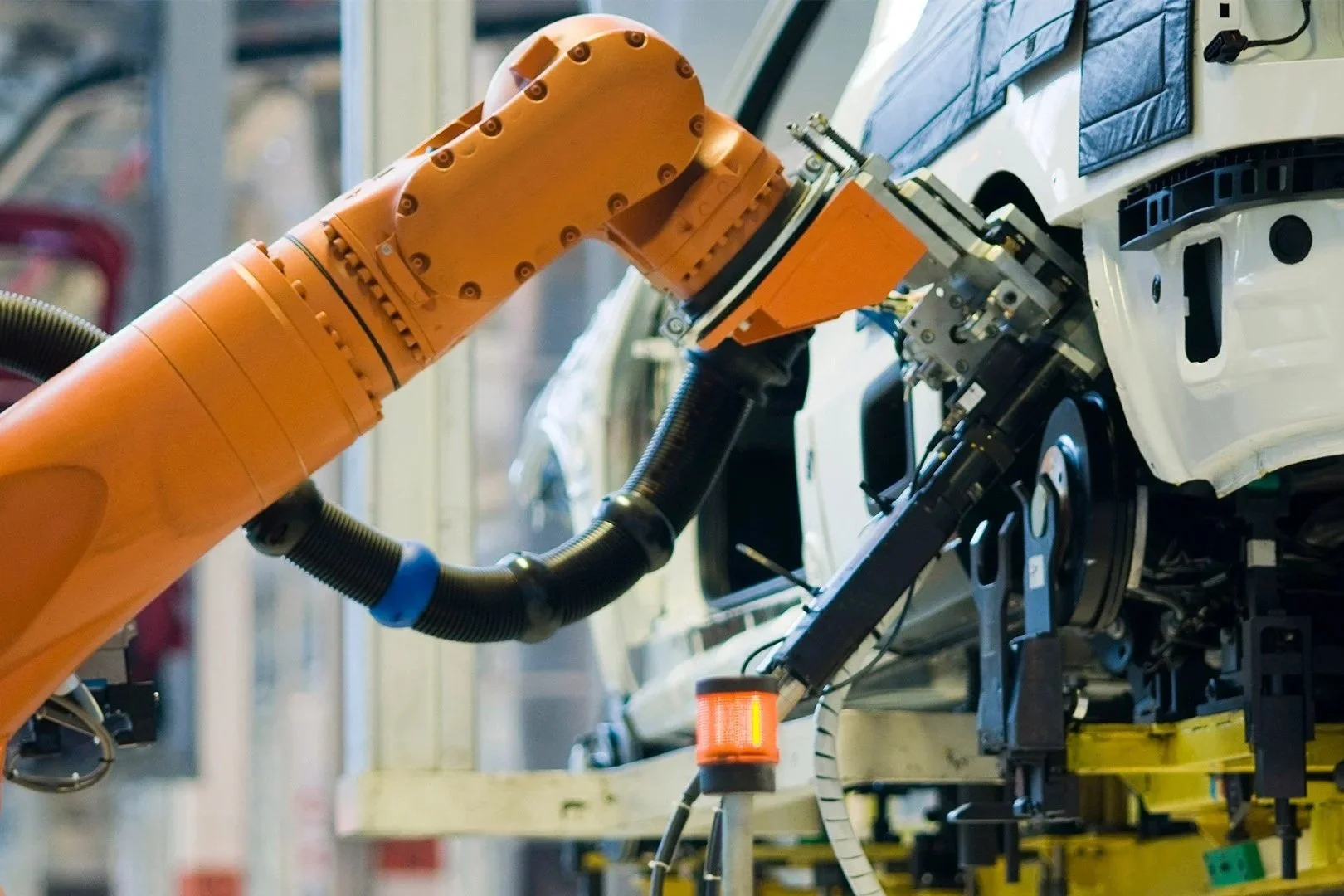 An orange robotic arm working on assembling or repairing a white vehicle in an automotive manufacturing plant.