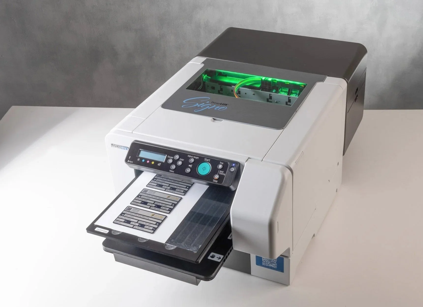 The Printolux Signo positioned on a white surface against a gray background. It is primarily white with a black and gray top section, and features a digital display panel and a tray with label plates.