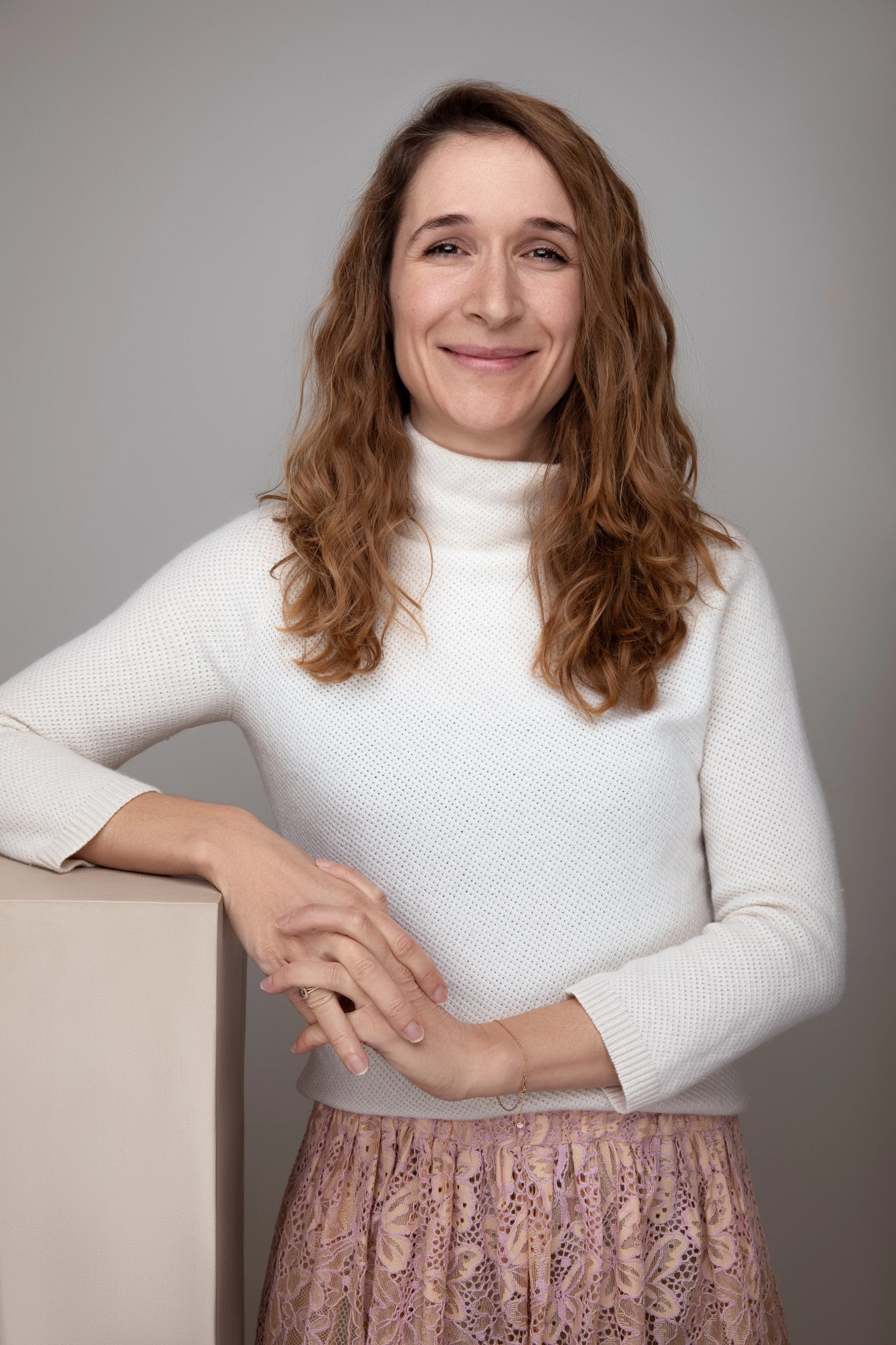 Et smilende kvindemodel iført en hvid sweater og en lyserød blonde nederdel, der står ved en væg.