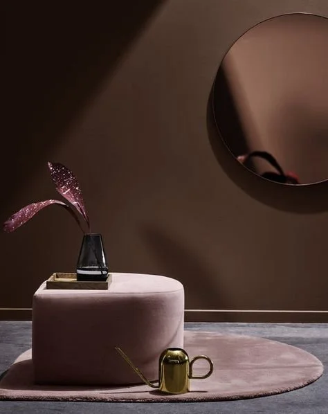 En rund, beige puffestol med en vovseplante i en sort vase og en guldtryk til vanding, placeret på en pink rund måtte. Bagved er en beige væg med en stor rund spejl og en del af en mørk væg.