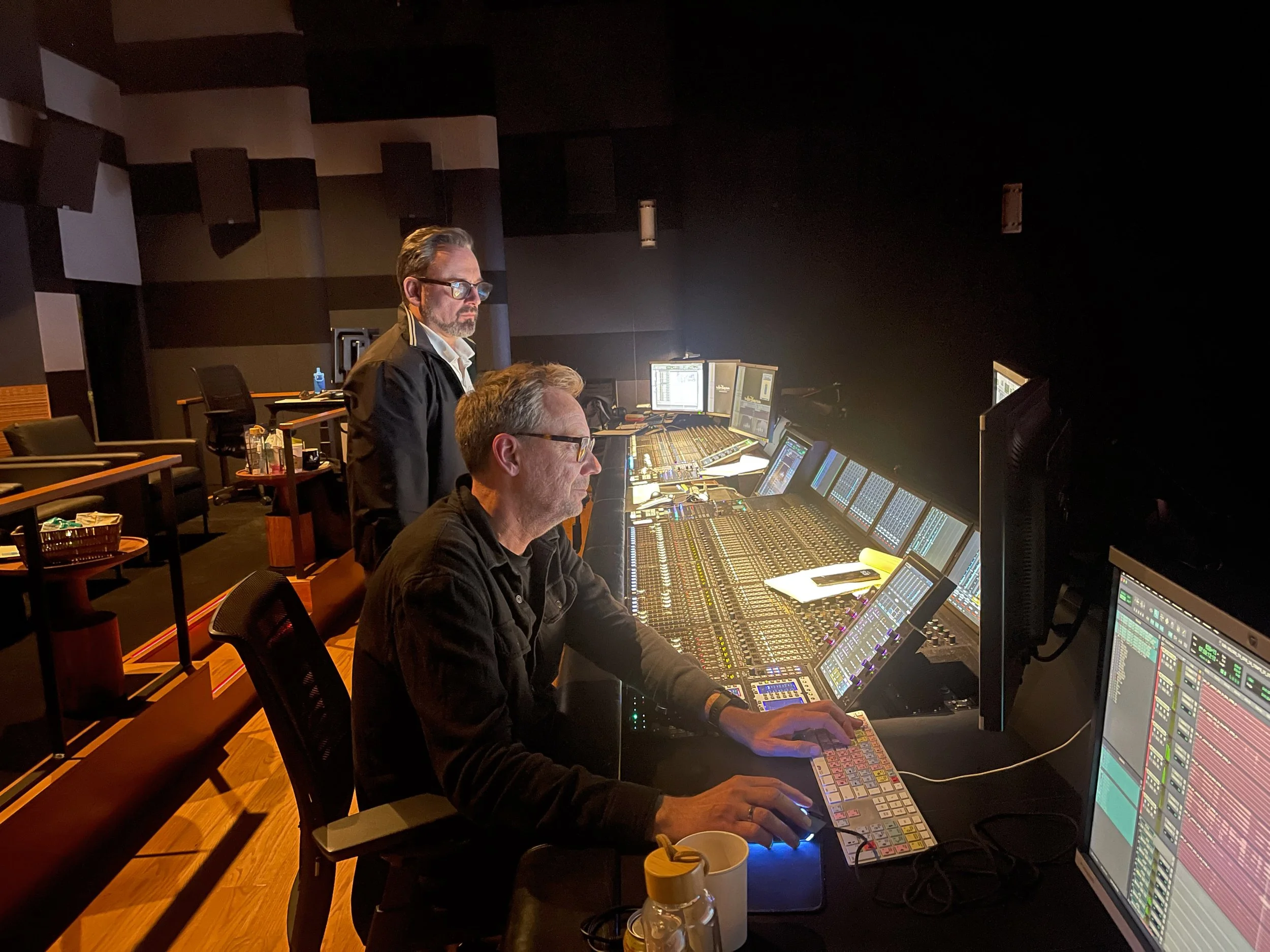Two men are working at a large audio mixing console in a recording studio, with multiple monitors, sound equipment, and empty chairs in the background.