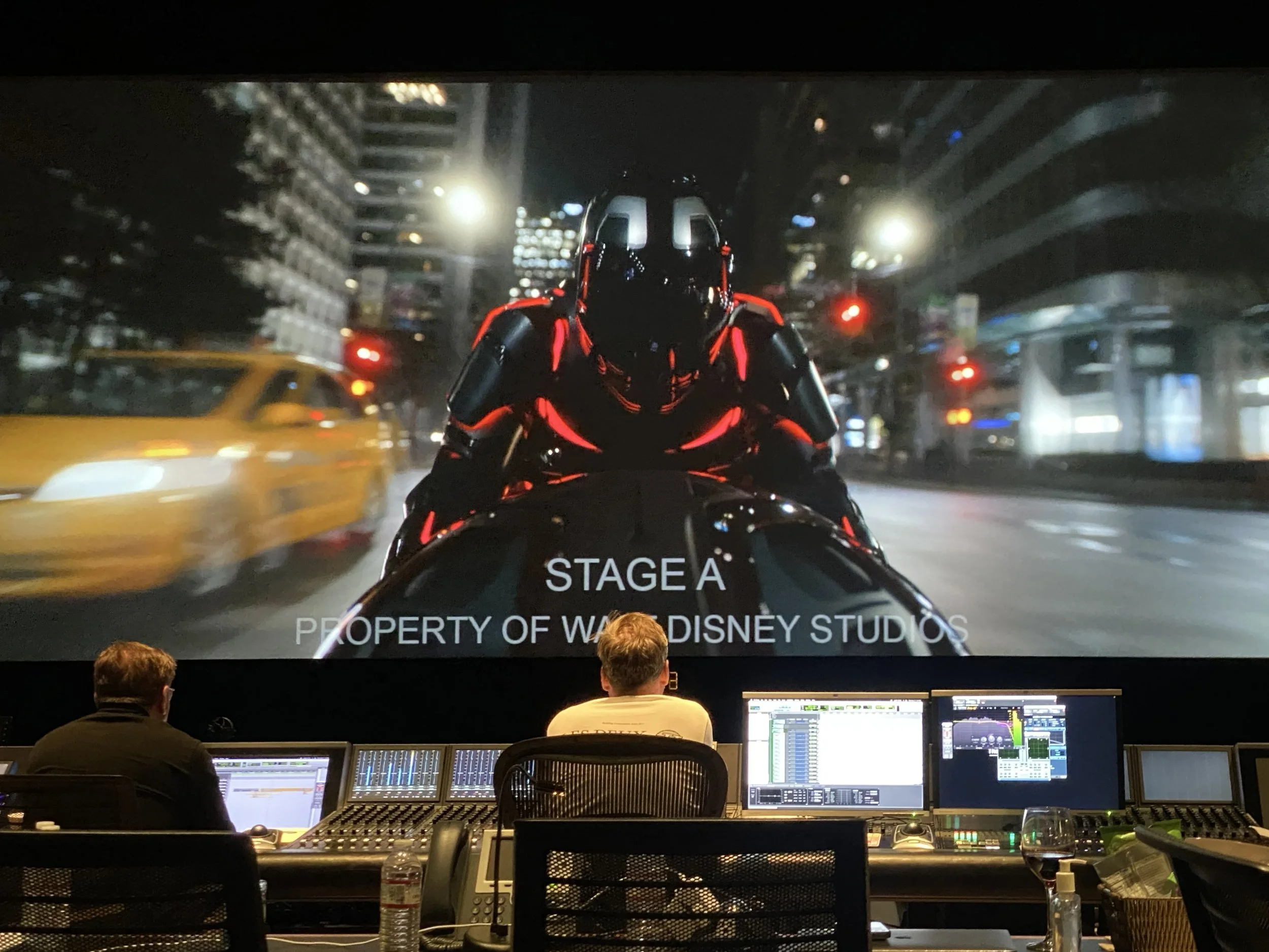 Sound editing studio with three people working at mixing consoles, watching a movie scene featuring a motorcycle rider on a city street at night, with the text "STAGE A PROPERTY OF WALT DISNEY STUDIOS" on the screen.