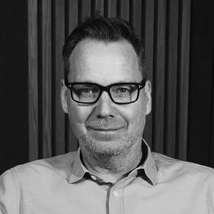 Black and white portrait of a man with glasses, short dark hair, wearing a light-colored button-up shirt, standing in front of a wooden panel background.