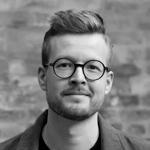 Black and white photo of a young man with glasses, short hair, and a beard, smiling, against a brick wall background.
