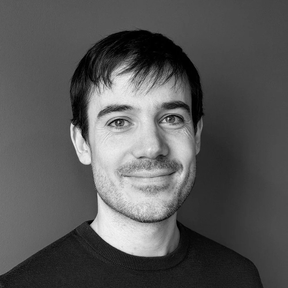 Black and white portrait of a smiling man with short dark hair and a light beard, wearing a dark shirt, against a solid dark background.