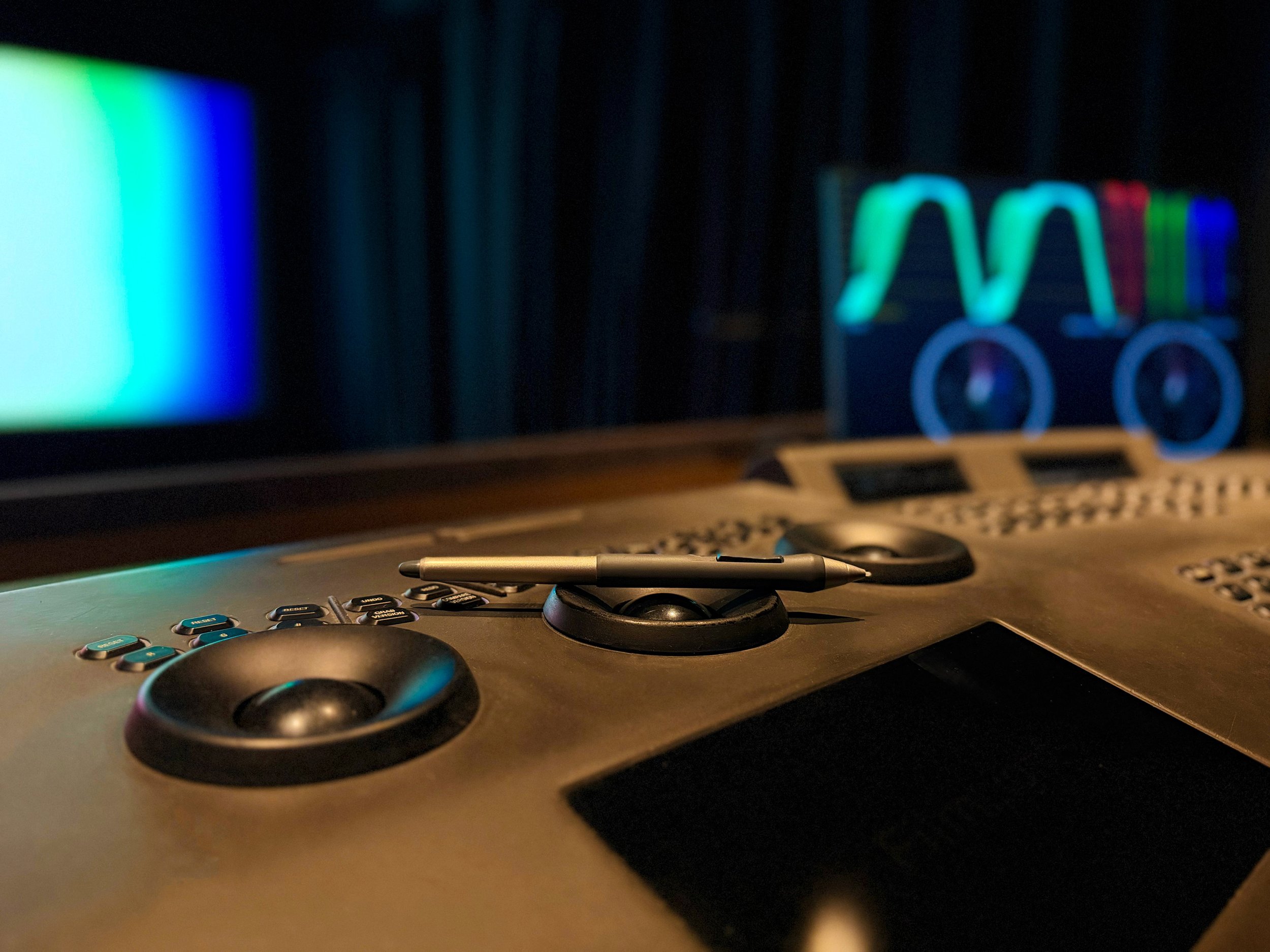 Close-up of a digital colorist console with a wacom pen resting on one of the knobs, in front of a computer monitor displaying colorful waveforms and a cinema screen in the background.