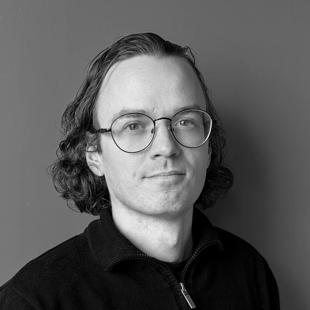 Black and white portrait of a man with shoulder-length wavy hair wearing glasses and a zip-up sweater, standing against a plain background.