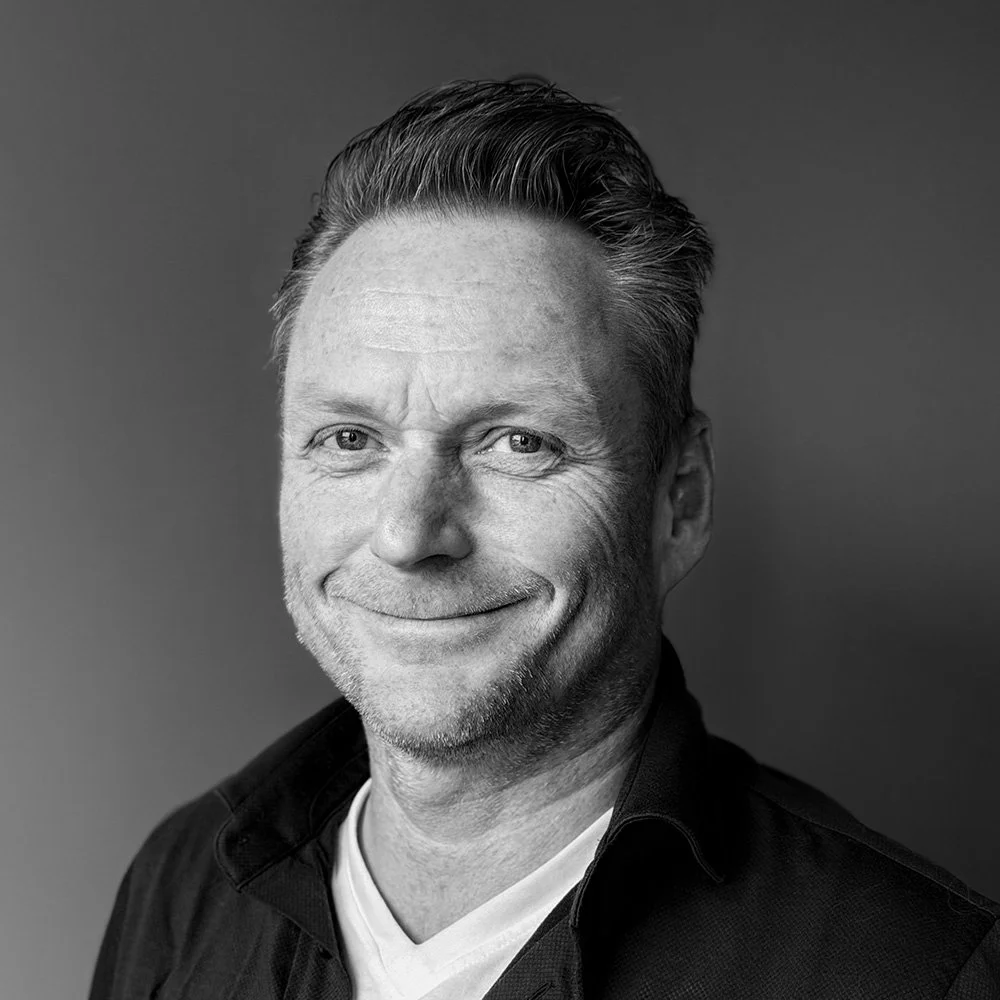 Black and white headshot of smiling middle-aged man with short, styled hair, wearing a dark jacket over a light shirt, against a plain background.