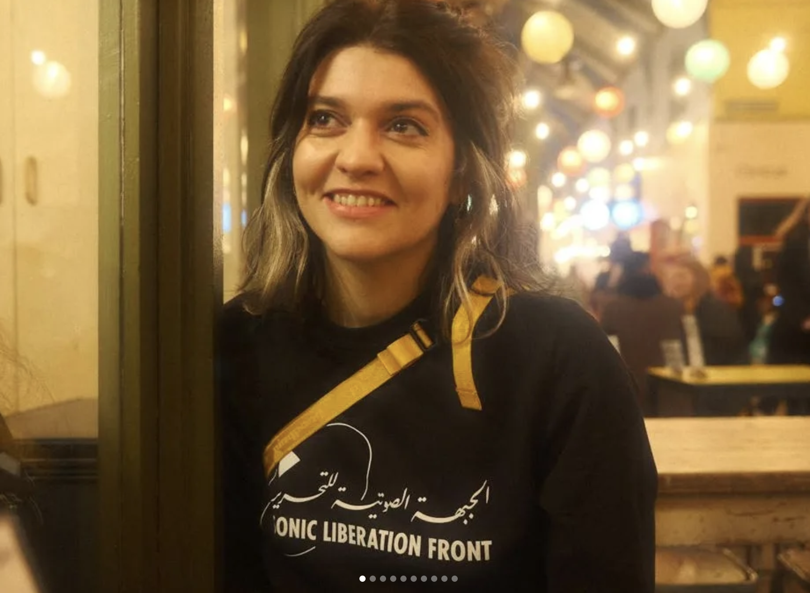 A woman with shoulder-length wavy hair smiling at night in a vibrant urban setting with colorful hanging lights and people seated at tables in the background. She wears a black shirt with white text and a yellow strap across her chest.
