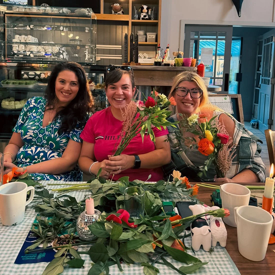 Three women sitting at a table with flower bouquets, cups, candles, and greenery in a cozy cafe with shelves of cups and plates in the background.