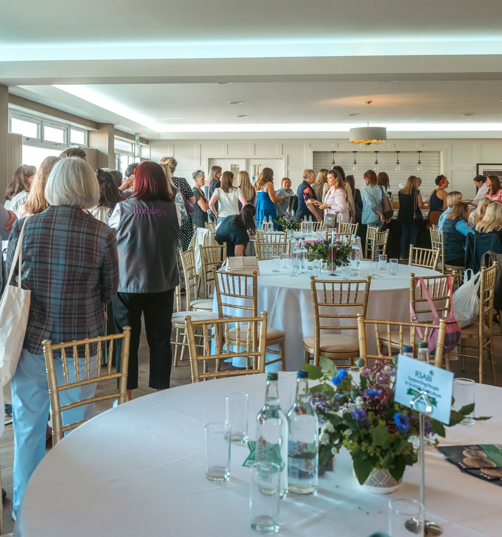 Women in Agriculture Royal highland show breakfast event with many women gathered, seated at decorated tables with floral centerpieces, in a well-lit room with large windows.