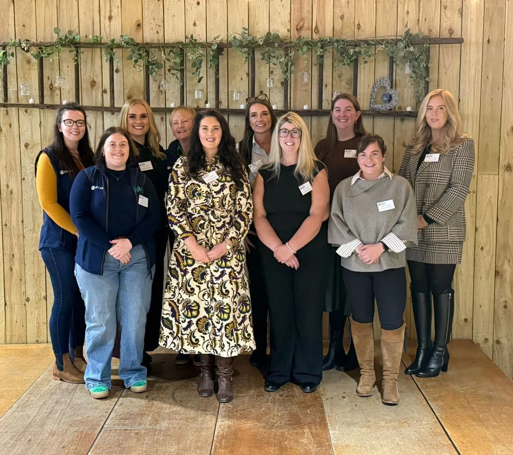 Women in agriculture 2026 committee photo. The Group of women are standing in front of a wooden wall decorated with greenery and hanging candles, smiling at the camera.