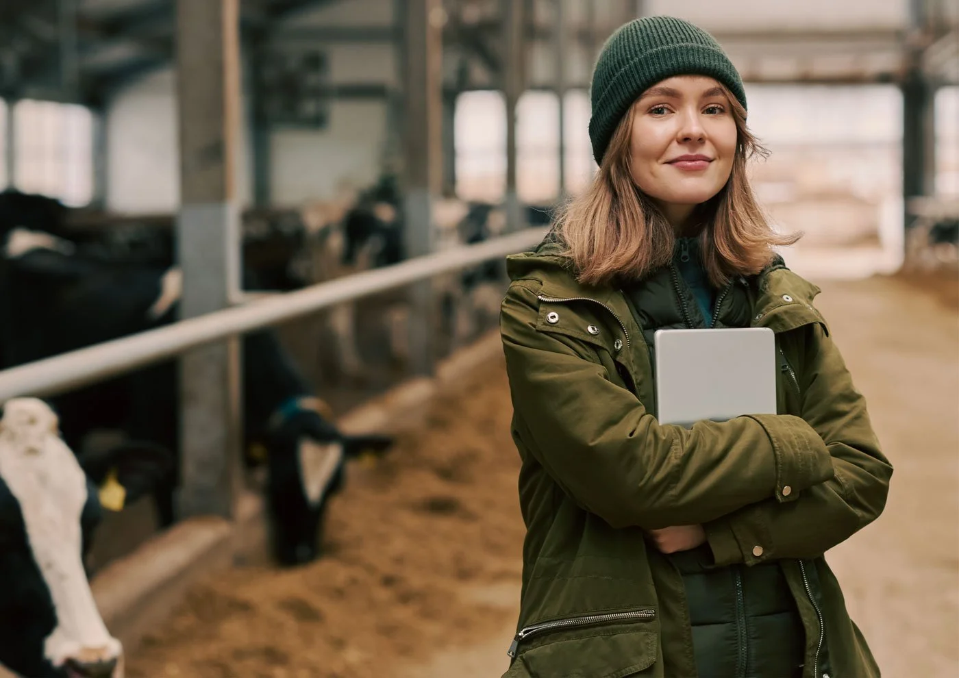 Young woman with light brown hair wearing a green beanie and jacket, holding a clipboard, standing in a barn with cows in background.
