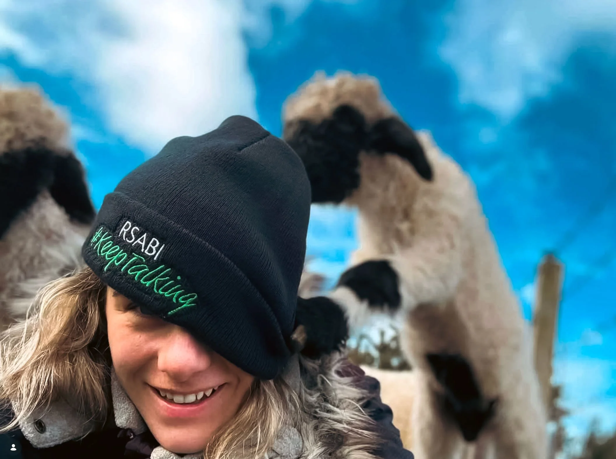 A woman smiling with her eyes obscured by her black beanie hat with green embroidery. She is next to a lamb who is jumping on her back and eating her hat.