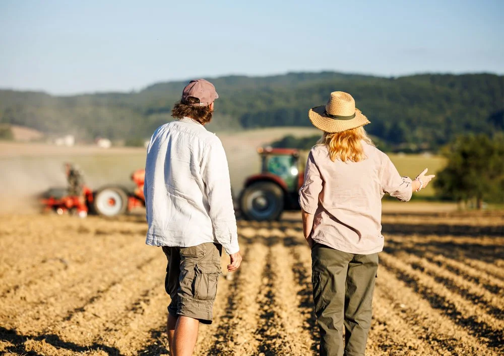 Two people walking in a farm field with a tractor in the background