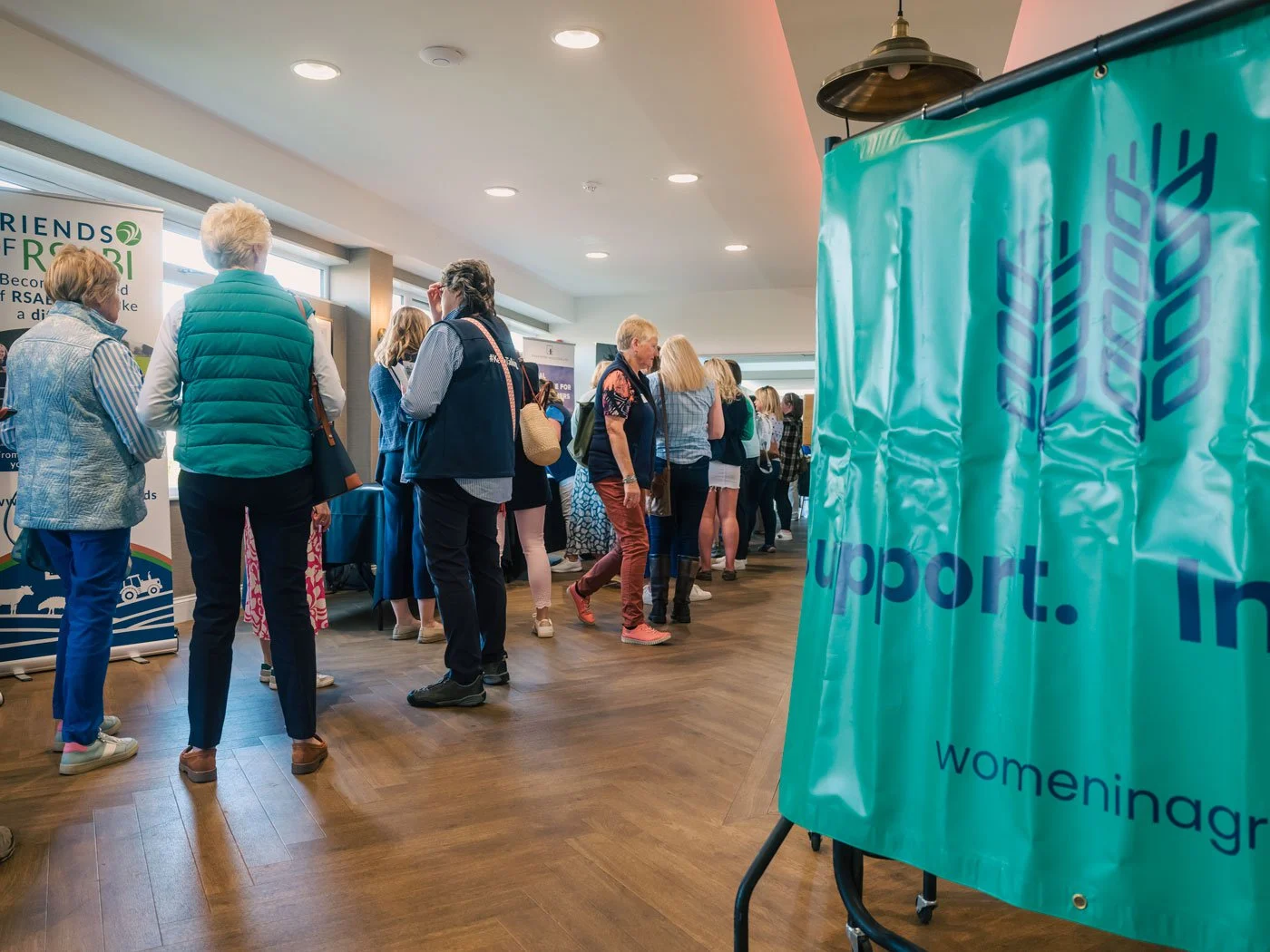 People standing in line at an the Women in Agriculture Royal Highland Show breakfast, with banners and booths visible, including a turquoise banner with arrows and the words 'support women in agriculture'.
