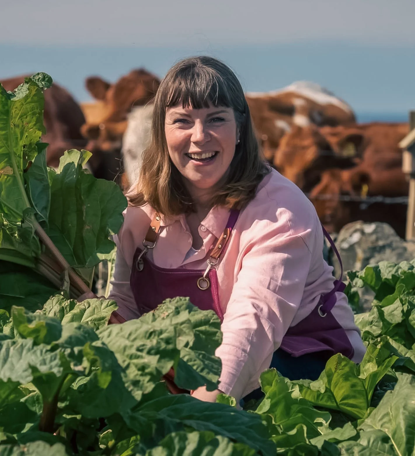 A woman smiling and working in a vegetable garden with leafy greens, with cows and a blue sky in the background.