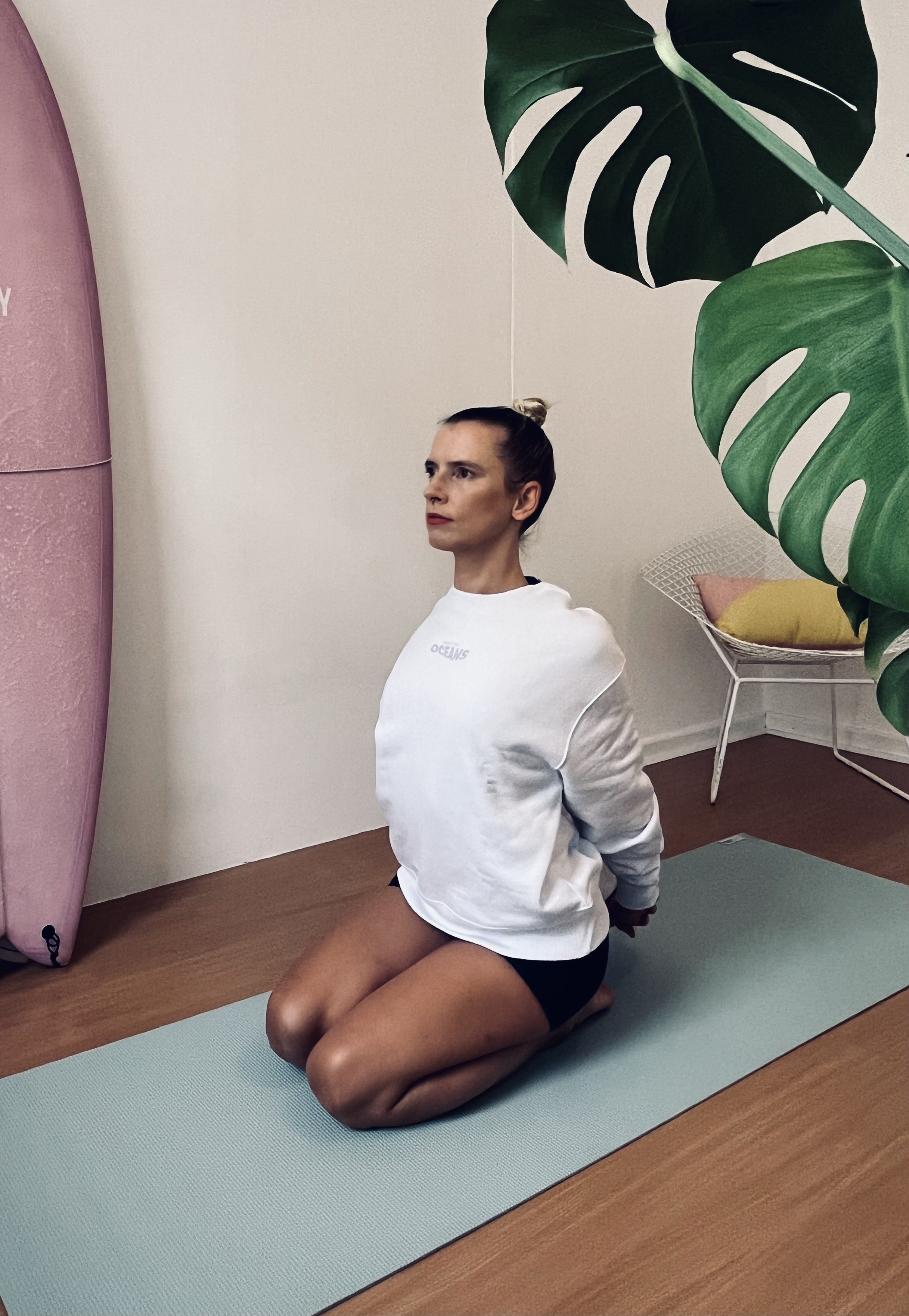 A woman practicing surf fitness on a light blue mat in a room with wooden floors, near a large pink surfboard, a white chair with a yellow cushion, and a large green monstera plant.
