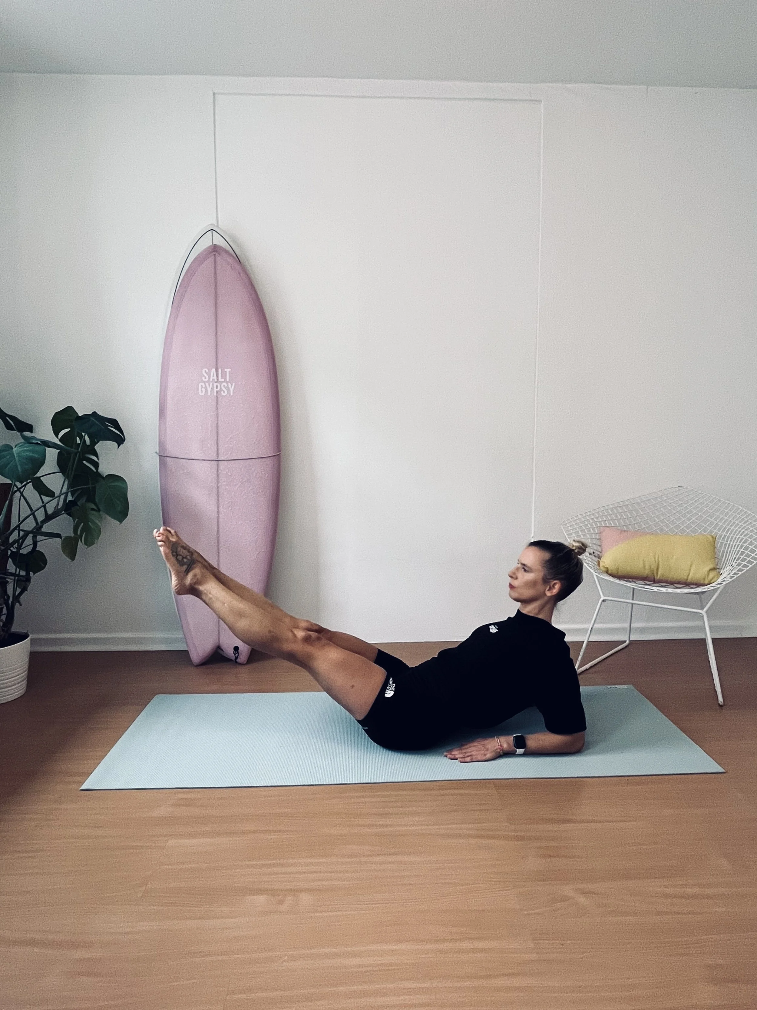 A woman doing leg lifts on a yoga mat in a room with a pink surfboard, a potted plant, and a white chair with a yellow pillow.