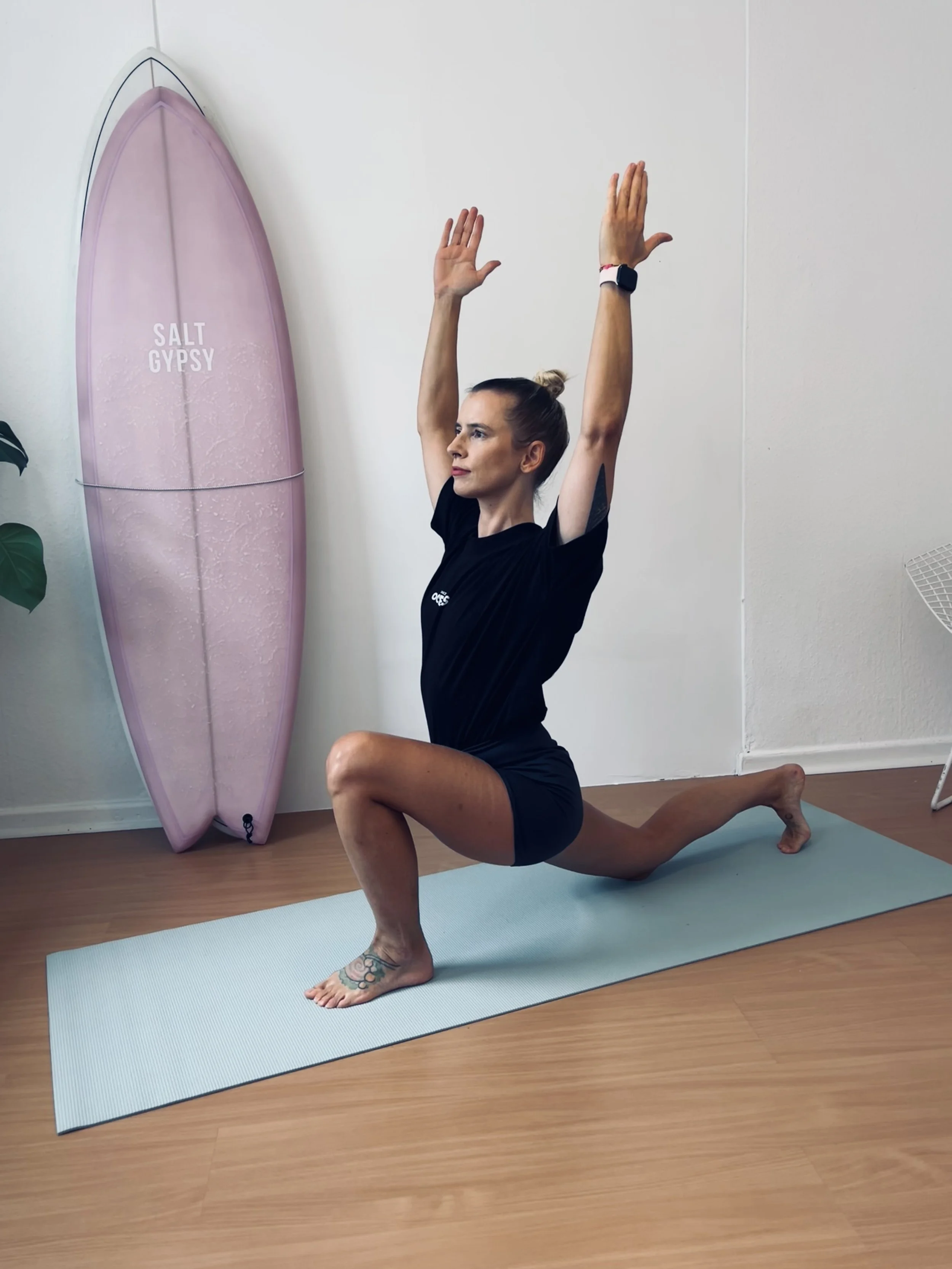 Woman practicing surf fitness in lunge pose in a room with a pink surfboard labeled 'Salt Gypsy' behind her, on a yoga mat on wood flooring.