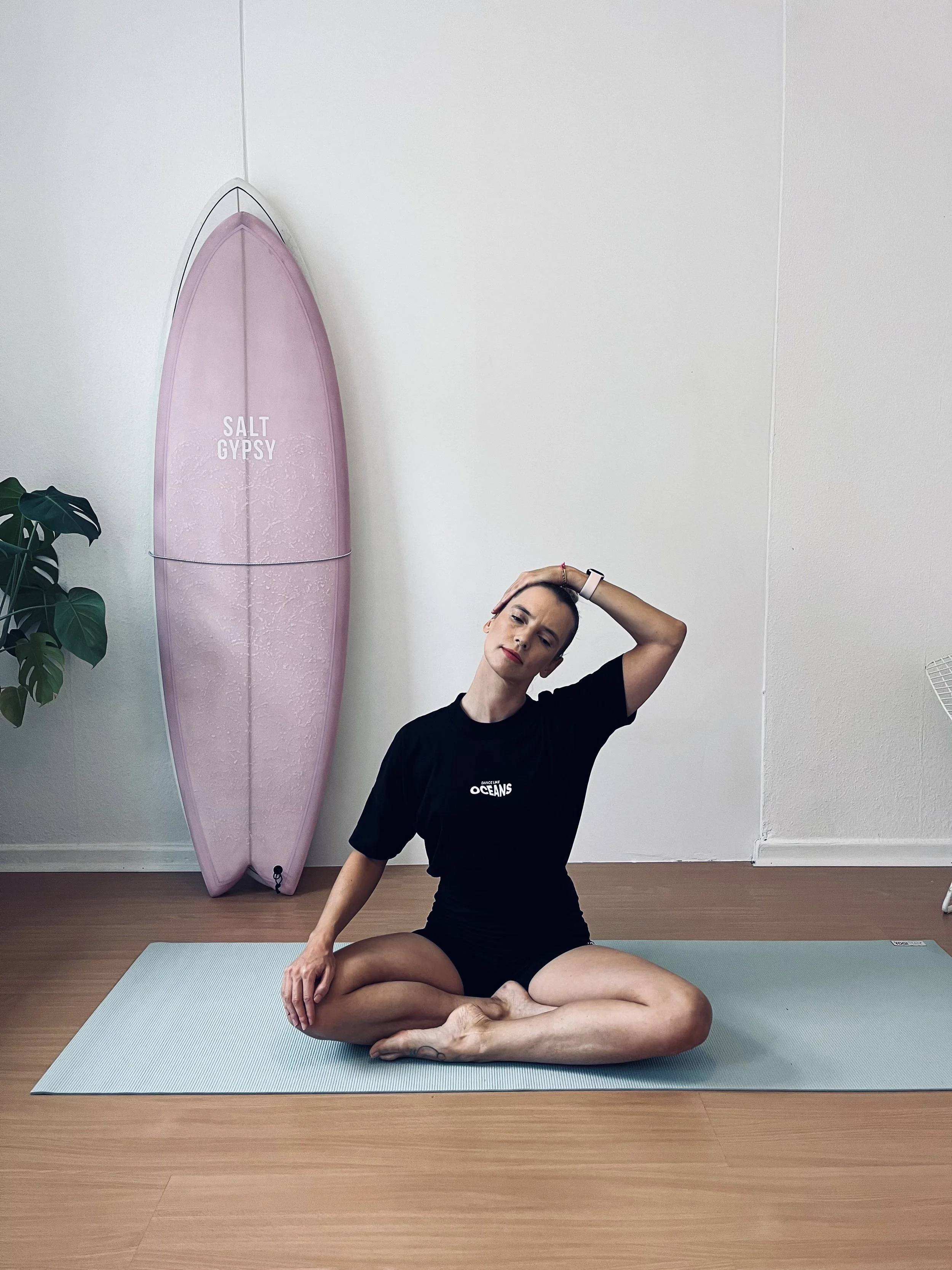 A woman practicing surf fitness on a light blue mat in a room with a pink surfboard and a plant in the background.